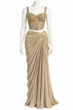 A floor-length beige evening gown displayed on a white mannequin against a plain white background. The dress features a heavily embellished corset-style bodice with gold embroidery and beading, paired with a draped, gathered skirt.