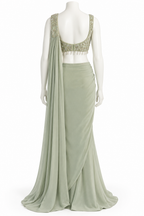 A sage green evening gown displayed on a white mannequin, featuring a sleeveless draped neckline with intricate gold embroidery and beadwork, a matching draped skirt, and a long flowing dupatta.