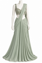 A sage green sari displayed on a white mannequin. The sari features a draped, flowing skirt and a matching draped dupatta, paired with a sleeveless, heavily embroidered light green and gold embellished blouse.