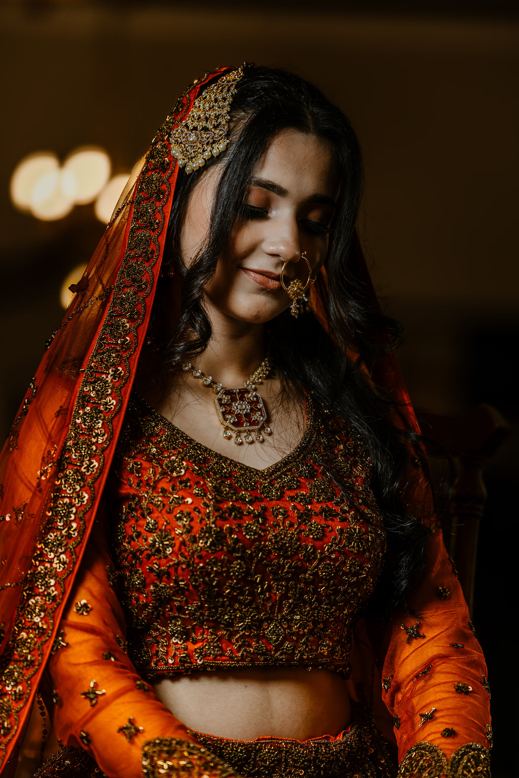 Woman in traditional orange and red embroidered outfit with jewelry, blurred lights in the background