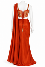 An orange-red traditional Indian outfit displayed on a white mannequin, featuring a draped skirt, a lace-up embroidered crop top, and a matching draped scarf.