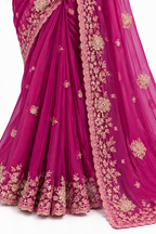 A close-up of a magenta pink georgette saree with intricate gold floral embroidery and a matching sheer dupatta draped over the skirt, set against a plain white background.