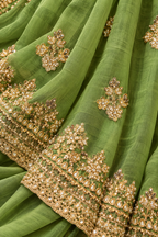 A close-up of a sage green fabric, likely a traditional dress material or dupatta, featuring intricate gold and silver mirror work embroidery along the borders and scattered floral motifs.