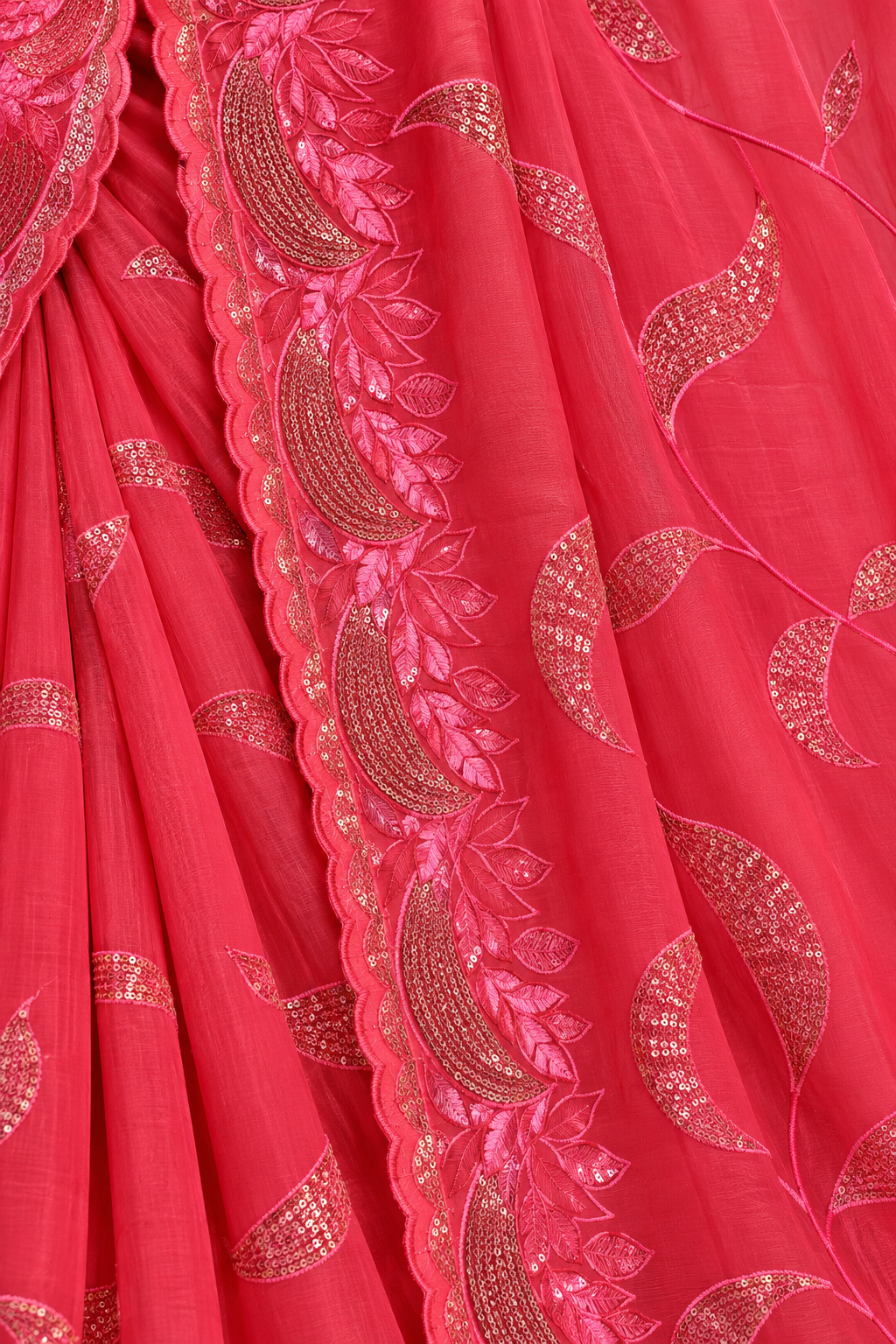 A close-up of a bright pinkish-red saree fabric with metallic gold floral and paisley embroidery and a scalloped border.