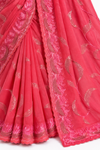 A close-up of a bright pink traditional fabric, likely a sari, featuring metallic gold floral and paisley embroidery and a scalloped pink border.