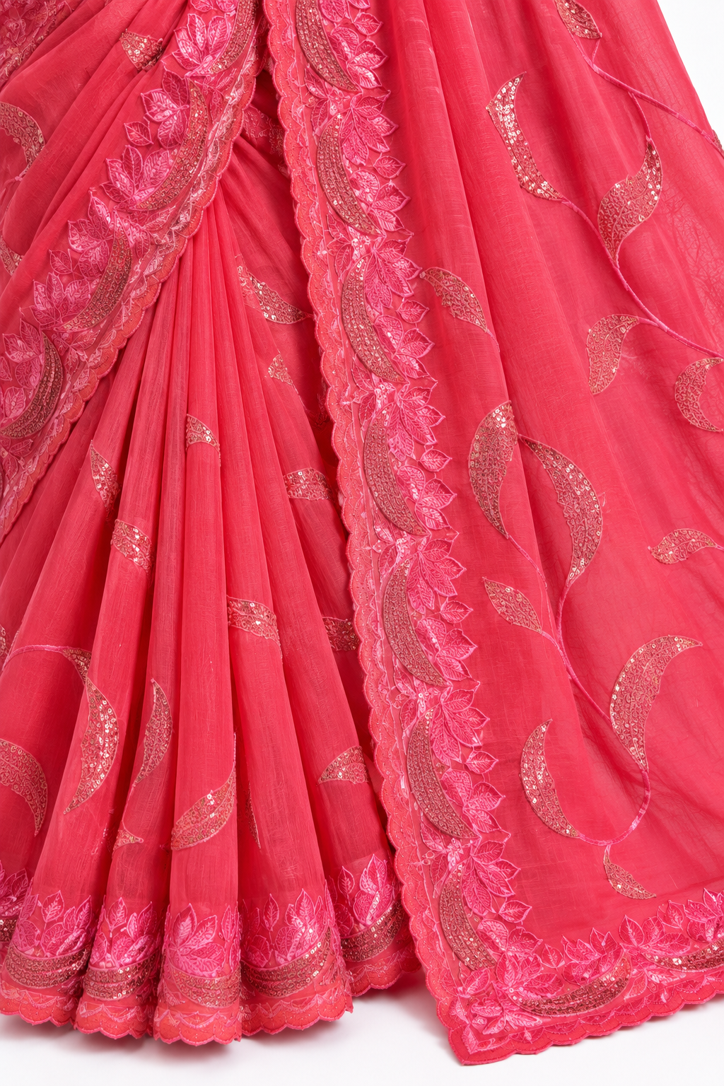 A close-up of a bright pink traditional fabric, likely a sari, featuring metallic gold floral and paisley embroidery and a scalloped pink border.