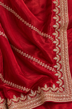 A close-up of a rich red fabric, likely a saree or dupatta, featuring detailed gold zari embroidery along the borders and draped elegantly.
