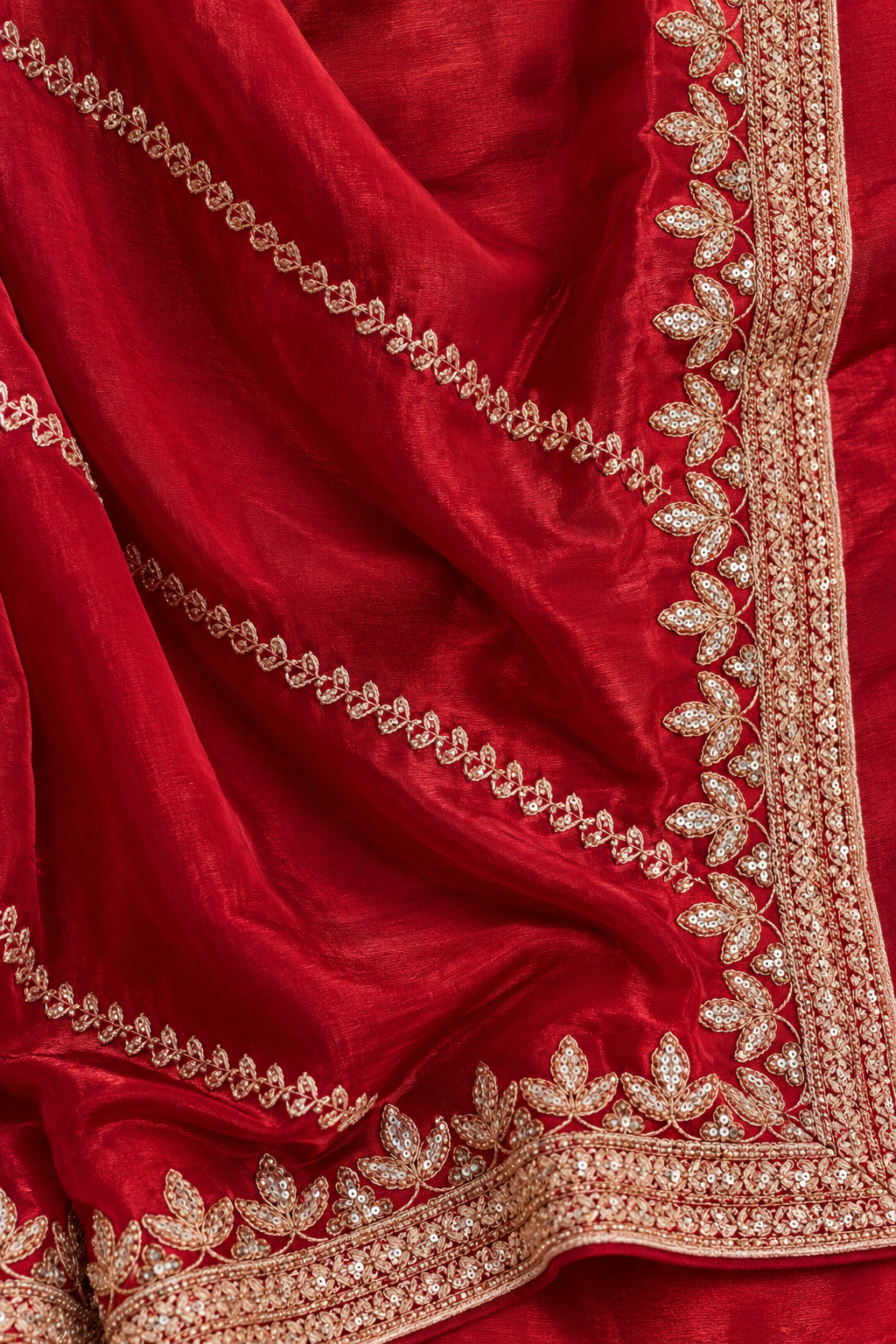 A close-up of a rich red fabric, likely a saree or dupatta, featuring detailed gold zari embroidery along the borders and draped elegantly.