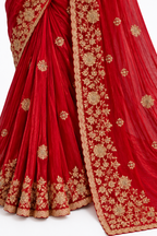 A close-up of a red draped fabric garment, likely a saree or lehenga, featuring intricate gold floral embroidery and a scalloped border, laid out against a white background.