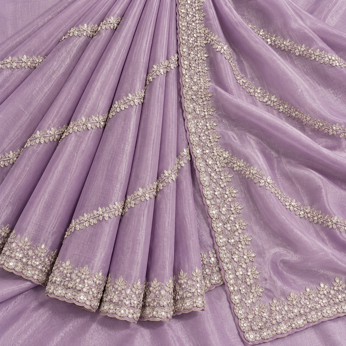 A close-up of a lavender purple fabric garment, likely a saree or dress, featuring detailed silver and white floral embroidery along the borders and draped diagonal stripes.