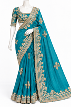 A teal blue saree with intricate gold floral embroidery and a wide gold border, displayed on a white mannequin against a plain white background.
