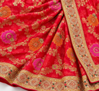 A close-up of a red Banarasi silk saree with intricate gold zari floral patterns and vibrant pink and orange embroidered motifs, featuring a detailed gold border.