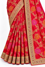 A close-up of a red sari with intricate gold, orange, and pink floral patterns and detailed gold borders, draped elegantly against a plain white background.