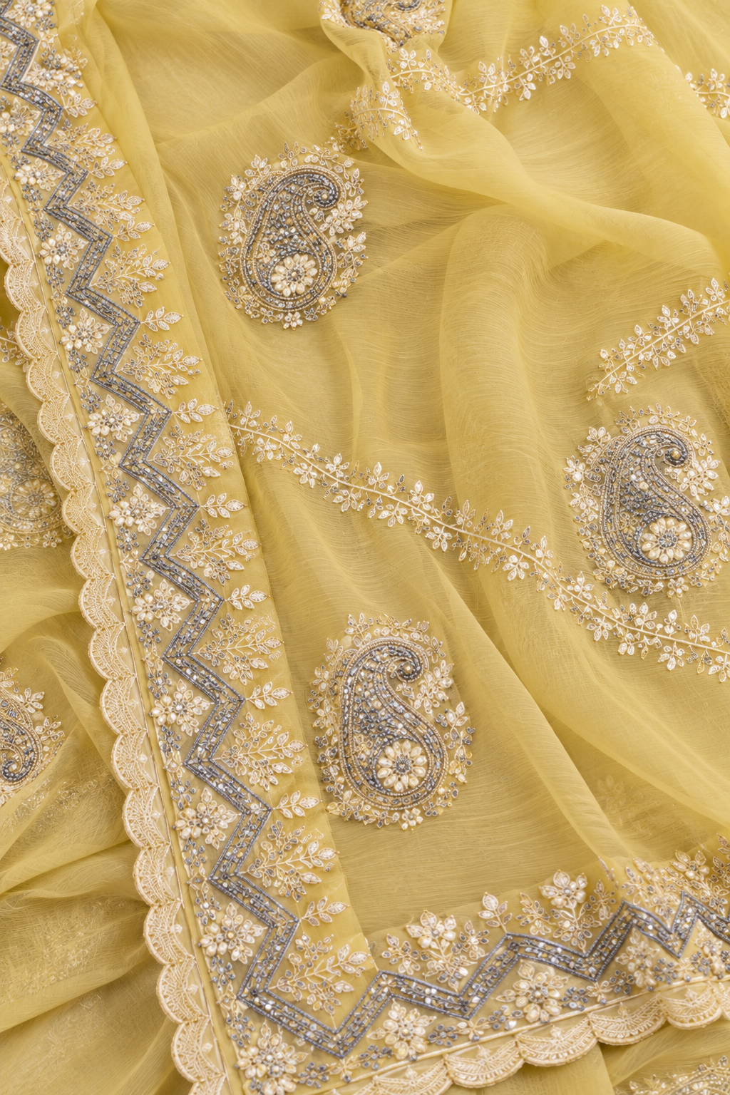 A close-up of a pale yellow sheer fabric with ornate silver and gold embroidered paisley motifs and scalloped borders.