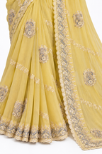 A close-up of a yellow saree with intricate silver and gold embroidery, featuring paisley motifs and a decorative scalloped border.