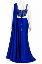 A royal blue formal gown displayed on a white mannequin, featuring a lace-up back bodice with beadwork, a matching draped scarf, and a flowing satin skirt.