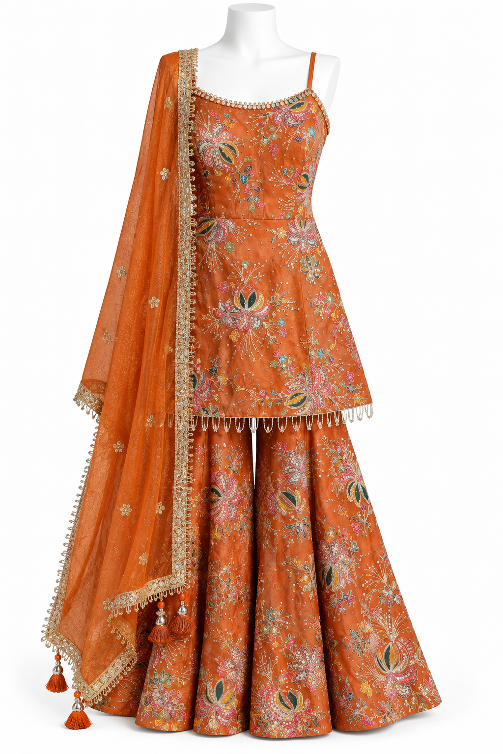 An orange embroidered traditional outfit displayed on a white mannequin against a white background. The set includes a sleeveless top with floral embroidery, matching flared pants, and a sheer dupatta with gold trim and tassels.