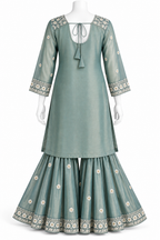 A sage green three-piece traditional Indian outfit displayed on a white mannequin, featuring a long kurta with a tie-neck and tassel, flared sharara pants, and matching embroidered pants.