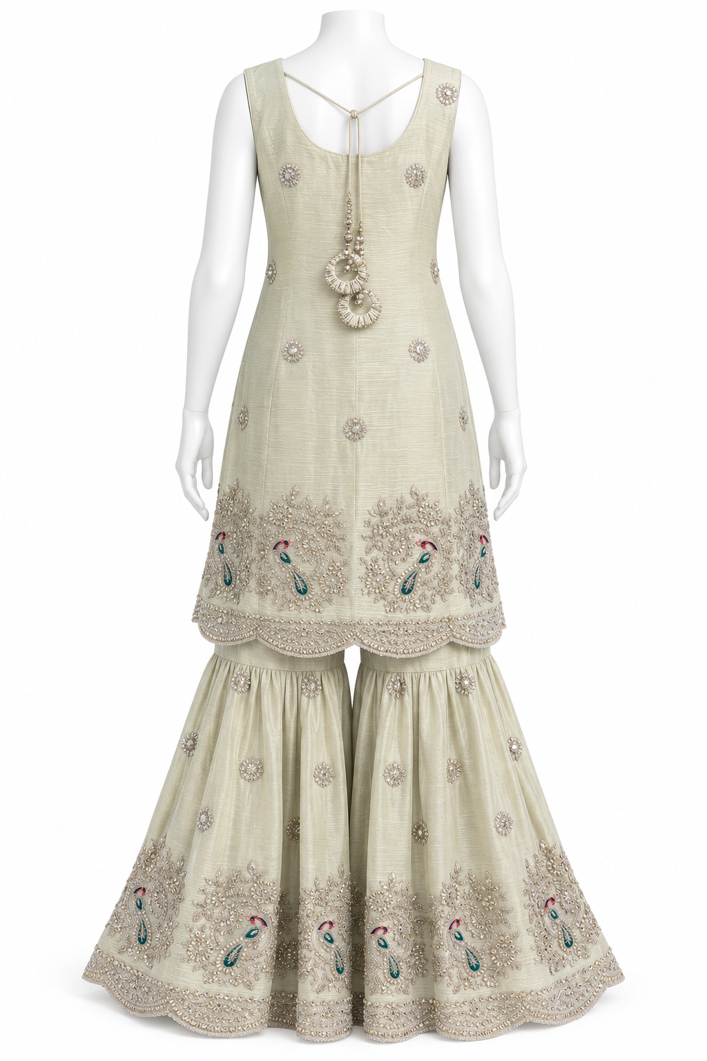 A sleeveless beige traditional outfit displayed on a white mannequin, featuring a long kurta with decorative embroidery and a matching flared sharara pant. The outfit has intricate metallic embroidery, beadwork, and peacock motif details near the hem and on the pants.