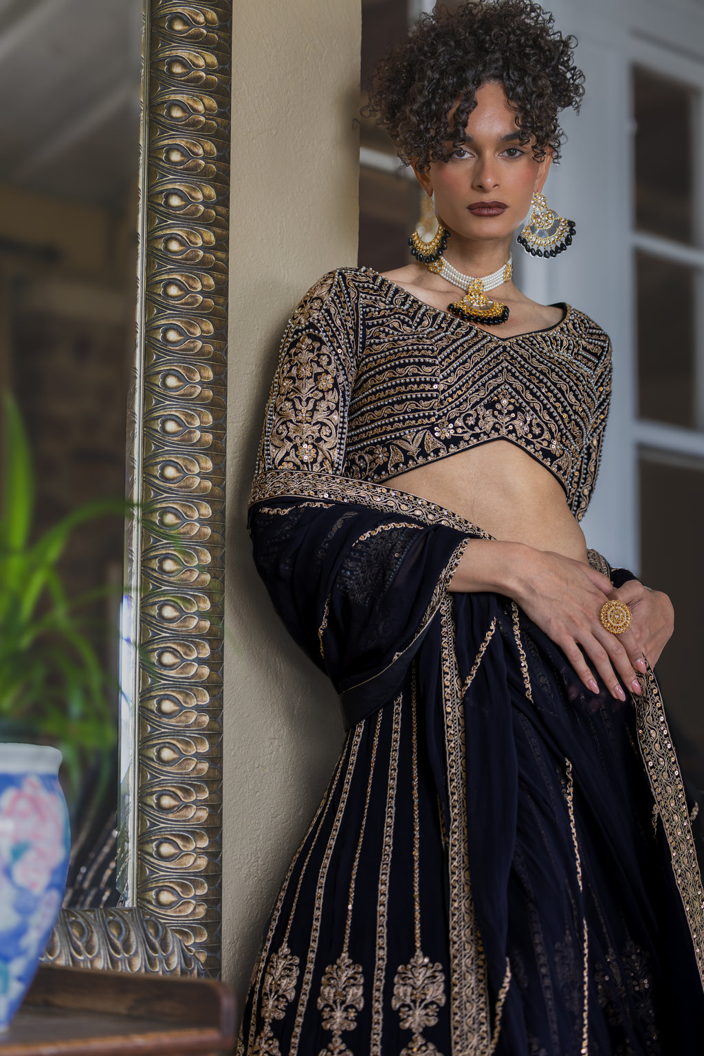 Woman in traditional black and gold outfit standing next to a decorative mirror.