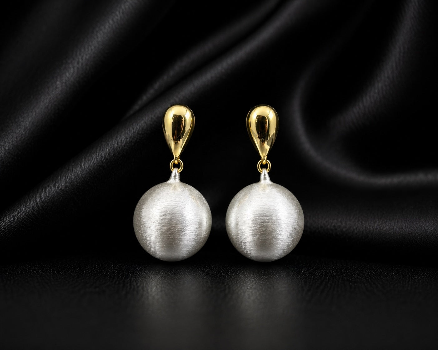 A pair of drop earrings featuring large, lustrous white pearl-like spheres suspended from polished gold teardrop-shaped hooks, displayed against a draped black satin fabric background.