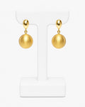 A pair of gold drop earrings with spherical dangling charms, displayed on a white T-shaped jewelry stand against a clean white background.