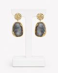 A pair of gold drop earrings with textured circular studs and irregular teardrop-shaped gray agate stones, displayed on a white T-stand against a white background.