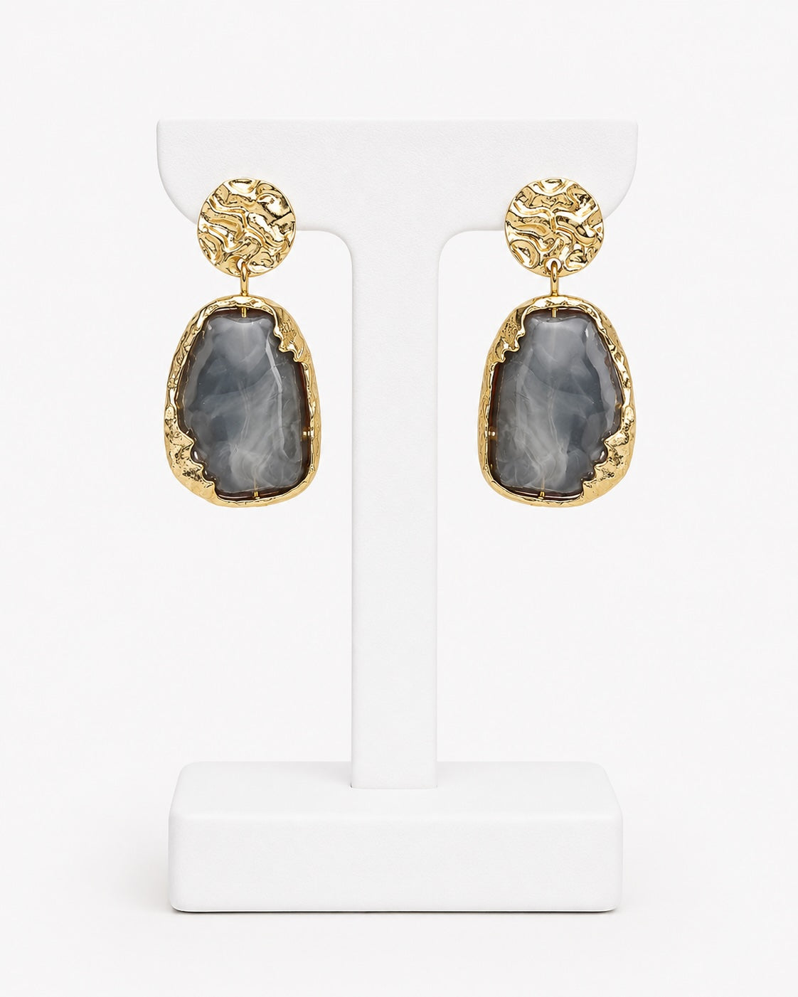 A pair of gold drop earrings with textured circular studs and irregular teardrop-shaped gray agate stones, displayed on a white T-stand against a white background.