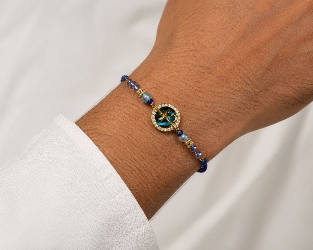 A rakhi worn on a person's wrist. The bracelet features deep blue beads, small gold spacer beads, and a central round gold medallion with a blue and green star motif surrounded by tiny white stones.