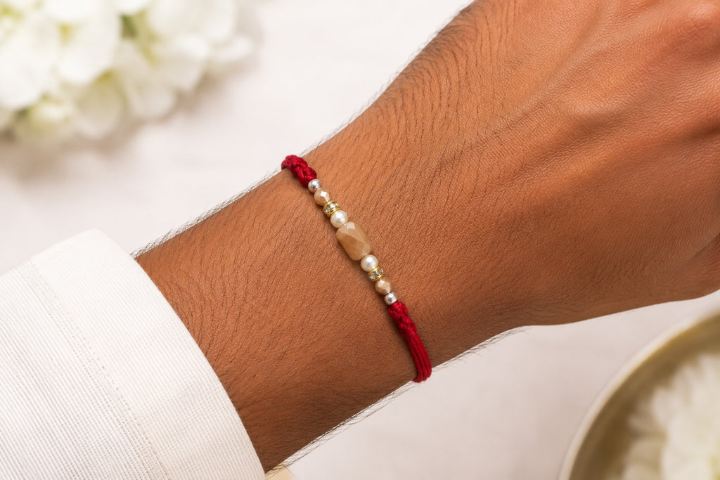A close-up of a person's wrist wearing a red cord bracelet with a central beige stone and small metallic beads, against a softly blurred light background.